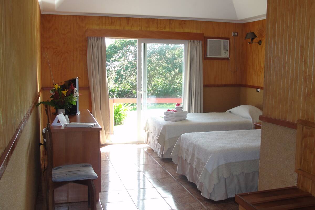 A hotel room with two beds, a desk with a chair, a small table with a vase of flowers, and a balcony with a view of greenery.