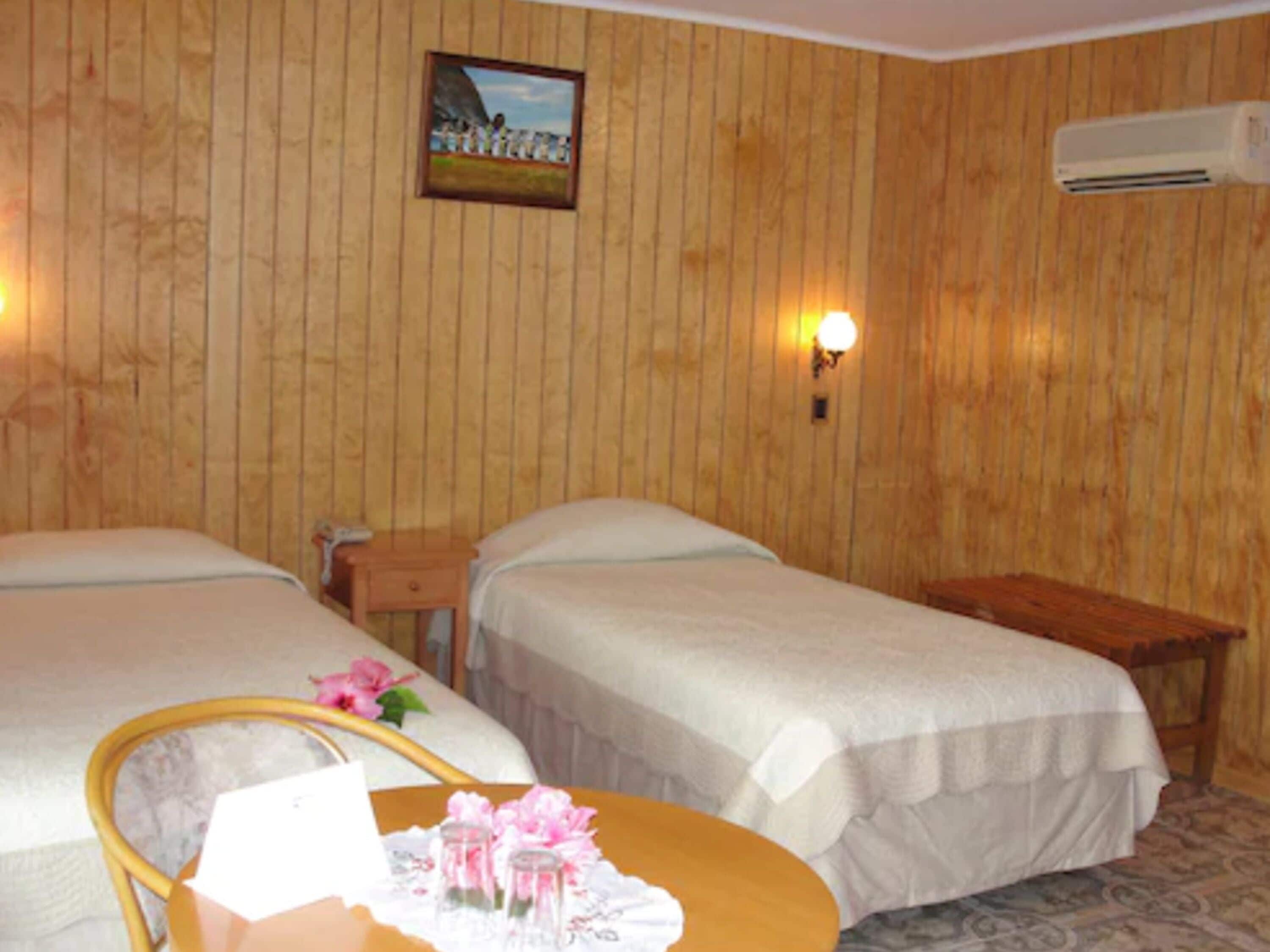 Habitación de hotel con dos camas, una pared de madera, un equipo de aire acondicionado y una pequeña mesa con un arreglo floral.
