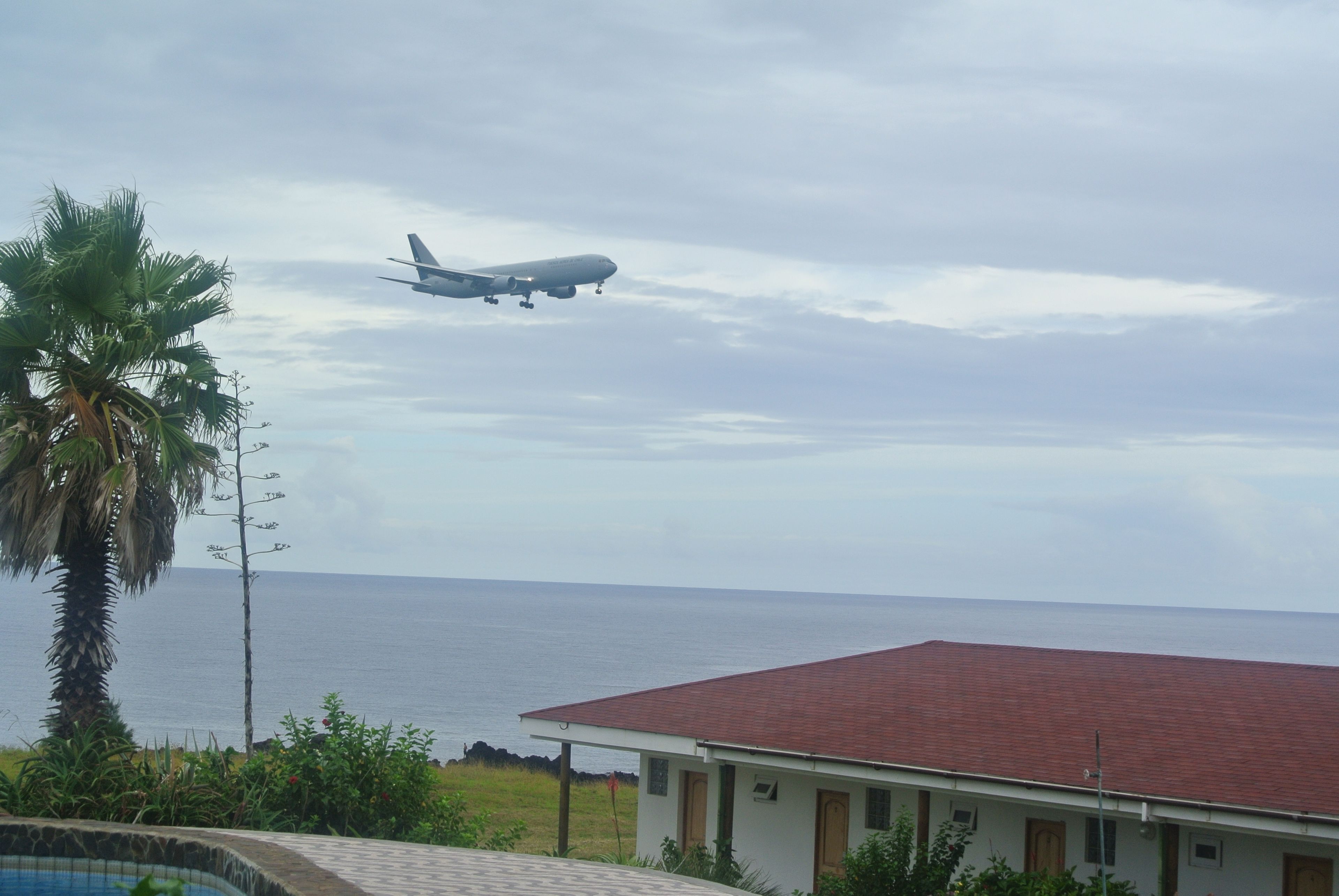 Um avião sobrevoa uma área costeira, com um edifício e palmeiras ao primeiro plano.