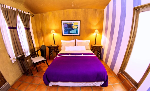 A hotel room with a large bed, two bedside tables, a chair, a painting on the wall, and curtains on the windows.