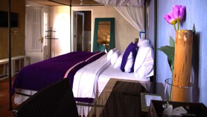 Un dormitorio con cama, mesita de noche, espejo, un jarrón de madera con una flor y ventana con cortinas.