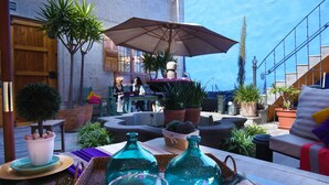 Un patio exterior con una mesa lista para comer, rodeado de plantas en macetas y un paraguas grande.