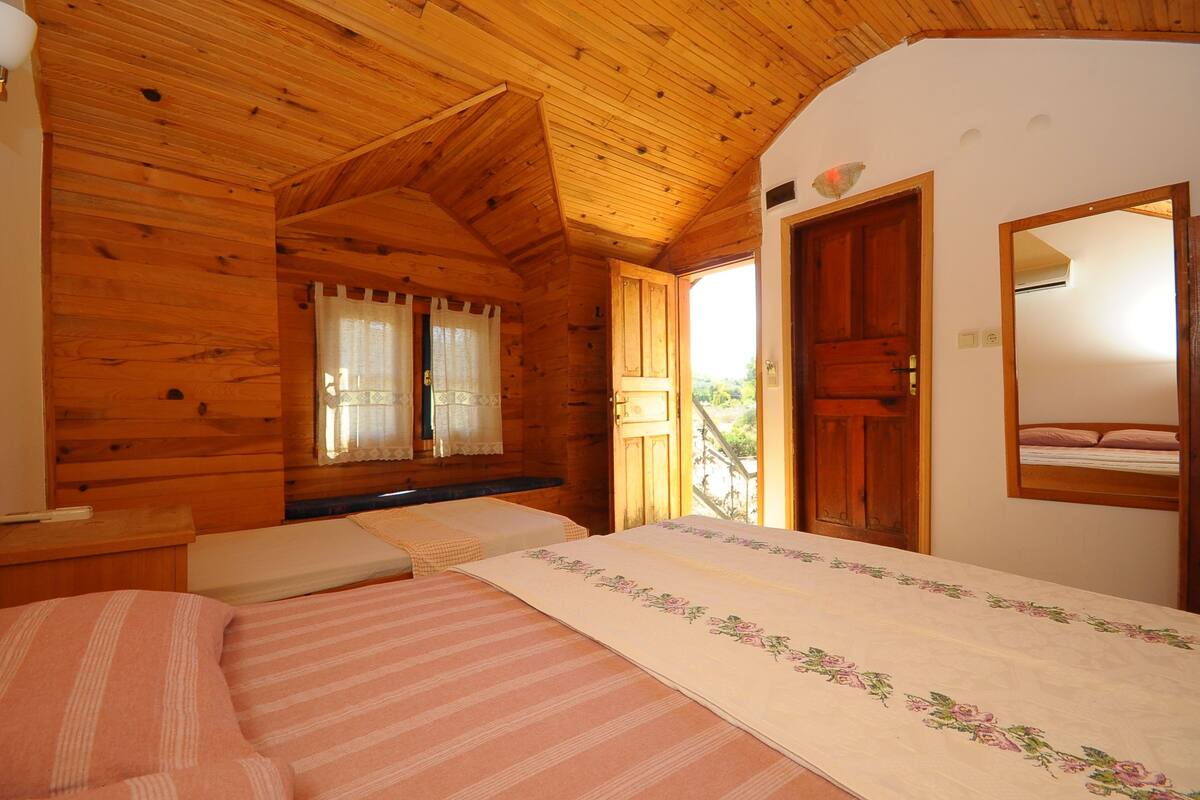 A wooden cabin bedroom with a bed, a nightstand, a window with curtains, and a door leading to another room.