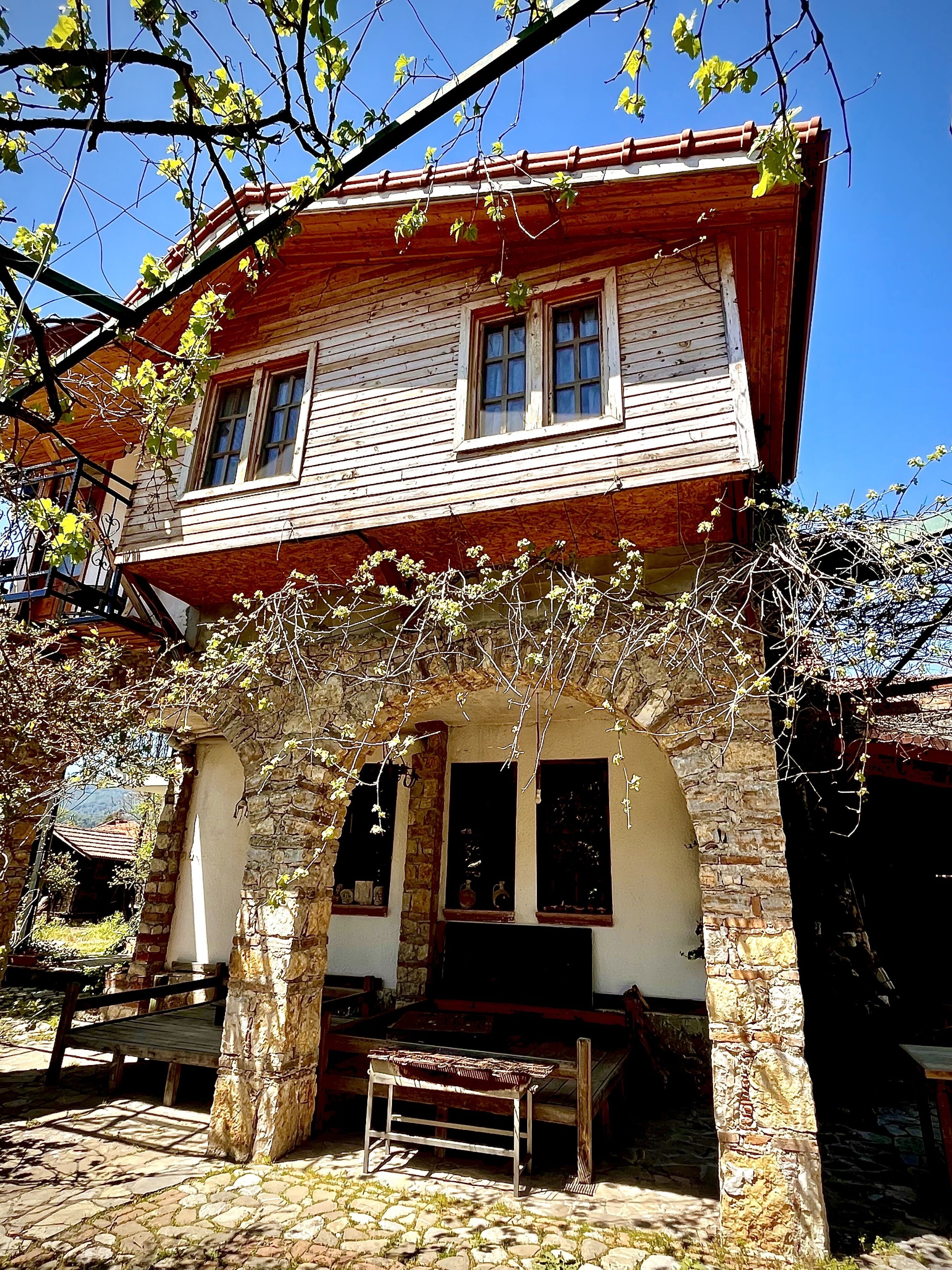 Una casa de madera con un arco de piedra en la entrada, un banco afuera y un cielo azul despejado.
