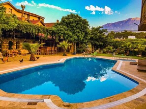 Una piscina con borde de piedra, rodeada de follaje y un edificio con una cruz en la parte superior.