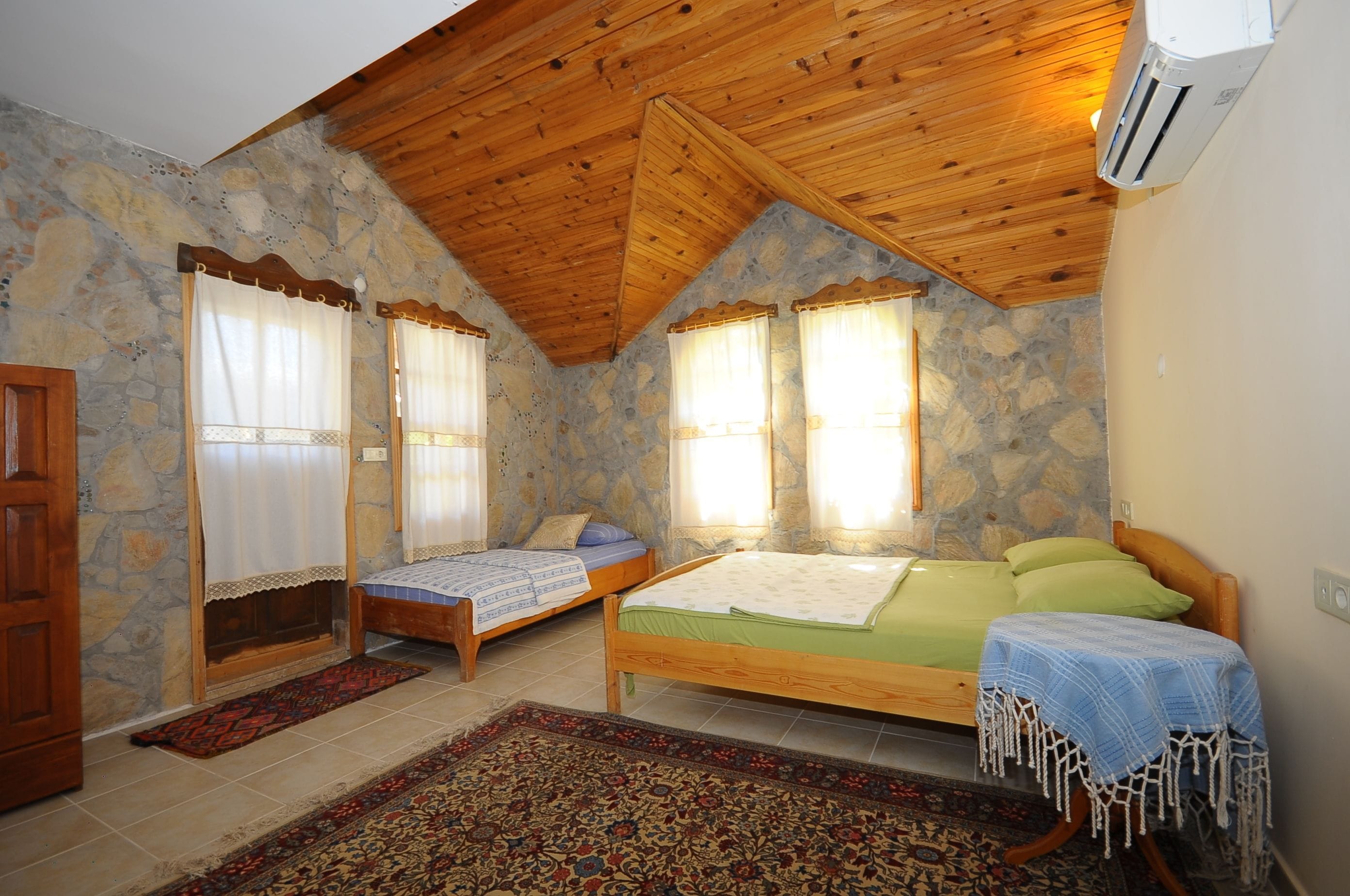 Una habitación con techo de madera inclinado, paredes de piedra, dos camas con ropa de cama verde y azul, y una alfombra estampada.