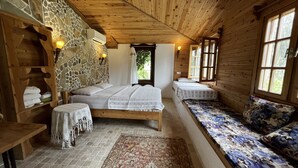 A cozy cabin room with a stone wall, wooden ceiling, a large bed, a bench with floral cushions, and a small table with a tablecloth.