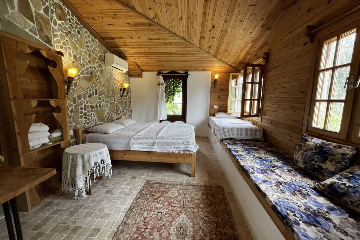 A cozy cabin room with a stone wall, wooden ceiling, a large bed, a bench with floral cushions, and a small table with a tablecloth.
