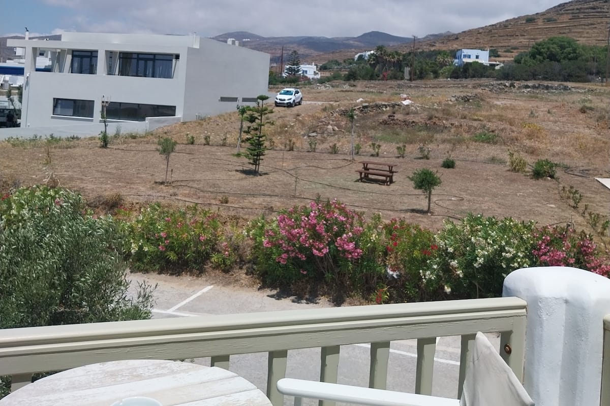 A balcony with a table and chairs, overlooking a dry landscape with sparse vegetation, a white building, and a distant mountain range under a clear sky.