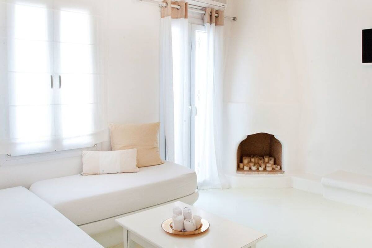 A minimalist living room with a white sofa, a small table with candles, a fireplace, and a TV mounted on the wall.