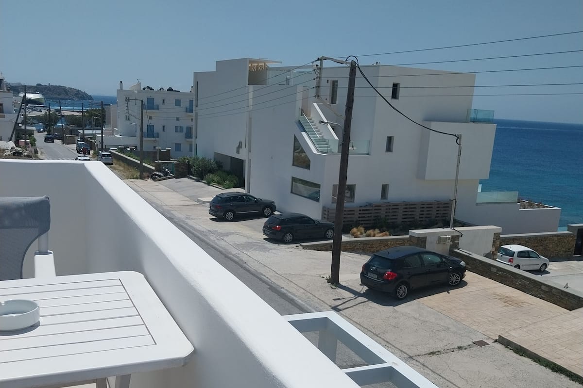 A balcony with a table and chairs, overlooking a street with parked cars and modern buildings, with a clear view of the sea in the background.