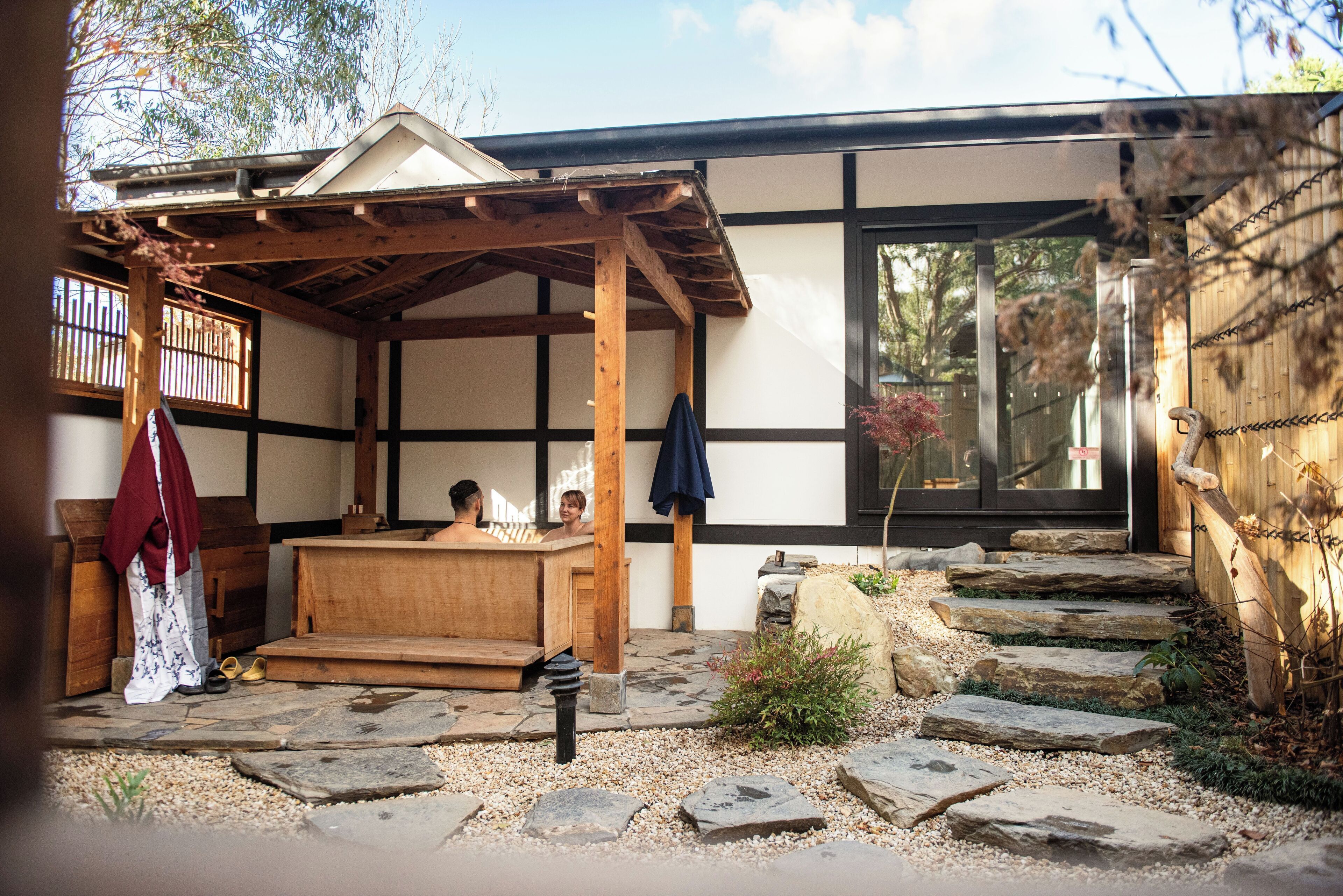 Un edificio tradicional japonés con suelo de esteras de tatami, estructura de madera y techo inclinado, y un sendero de piedra que conduce a la entrada.