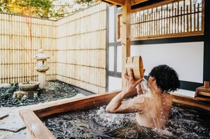 Una persona en un baño de aguas termales de madera, con una cerca de bambú y un farol de piedra al fondo.
