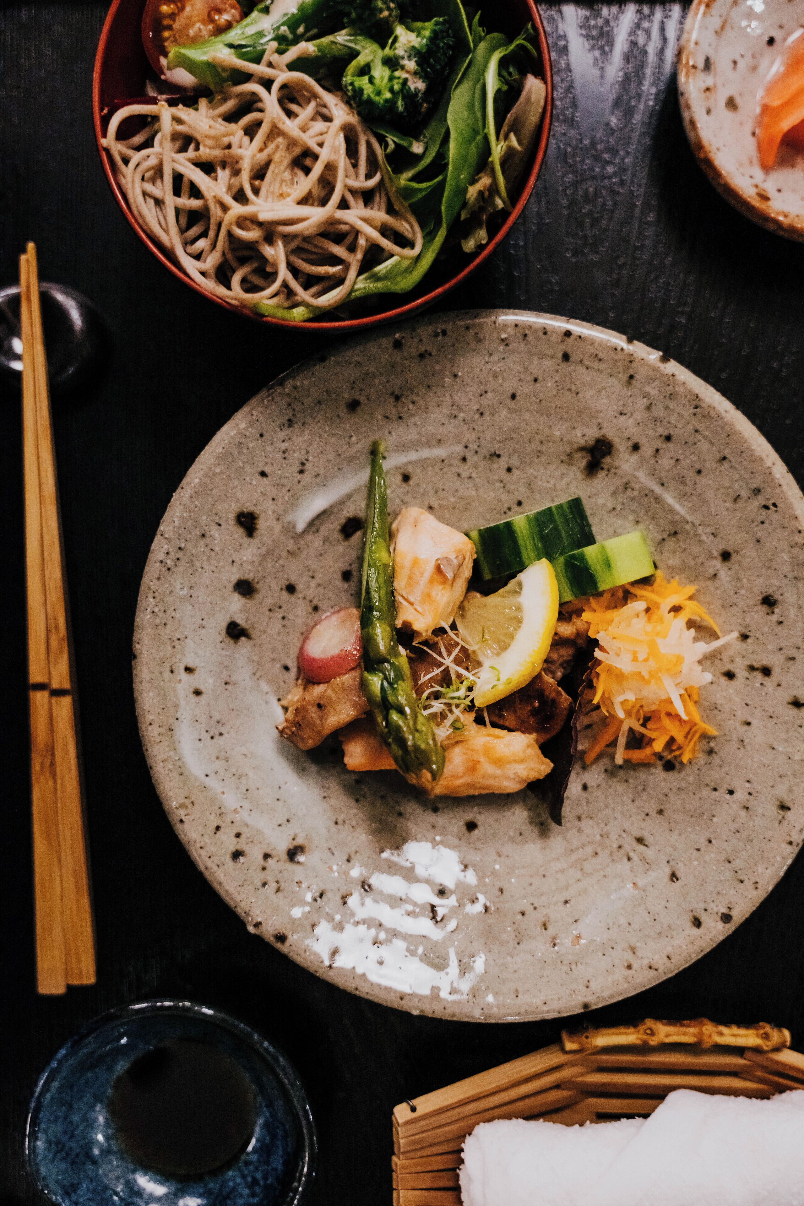 A table with a plate of food, including noodles, vegetables, and a slice of lemon.