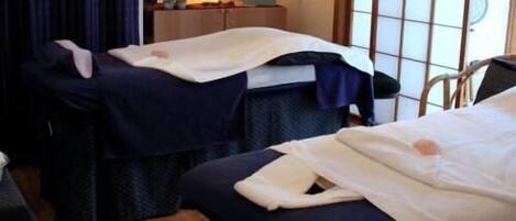 A room with two massage tables, a small desk, a chair, and a window with a view of the outside.
