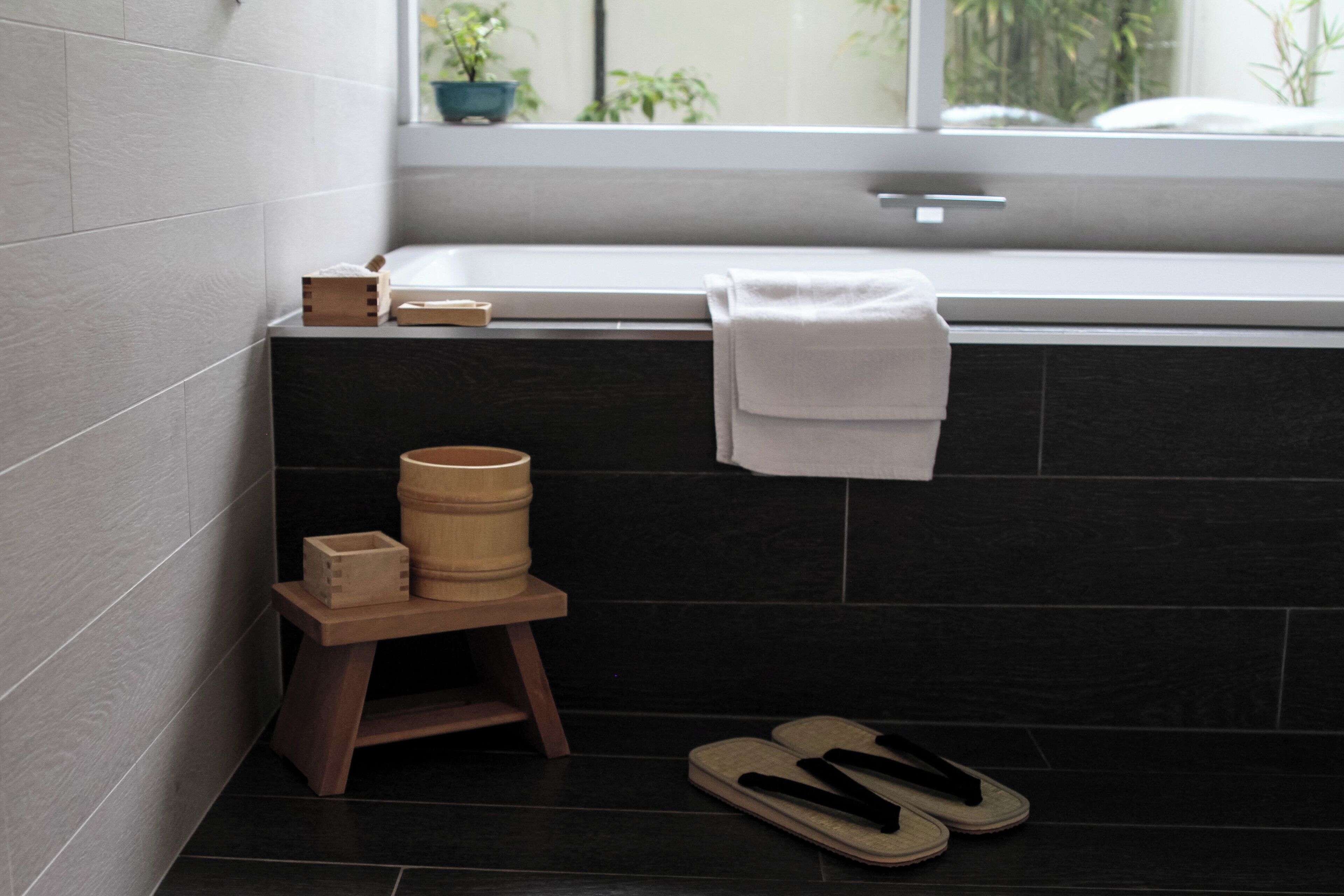 Un baño con una bañera, un taburete de madera con objetos encima y un par de pantuflas en el suelo.
