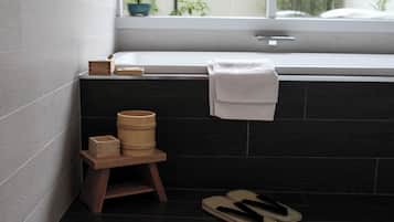 Un baño con una bañera, un taburete de madera con objetos encima y un par de pantuflas en el suelo.