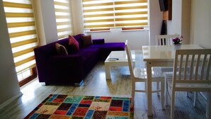 A living room with a purple sofa, a white table, and a white chair. There are windows with blinds, a TV mounted on the wall, and a colorful rug on the floor. - Ale Boutique Hotel (Konyaalti)