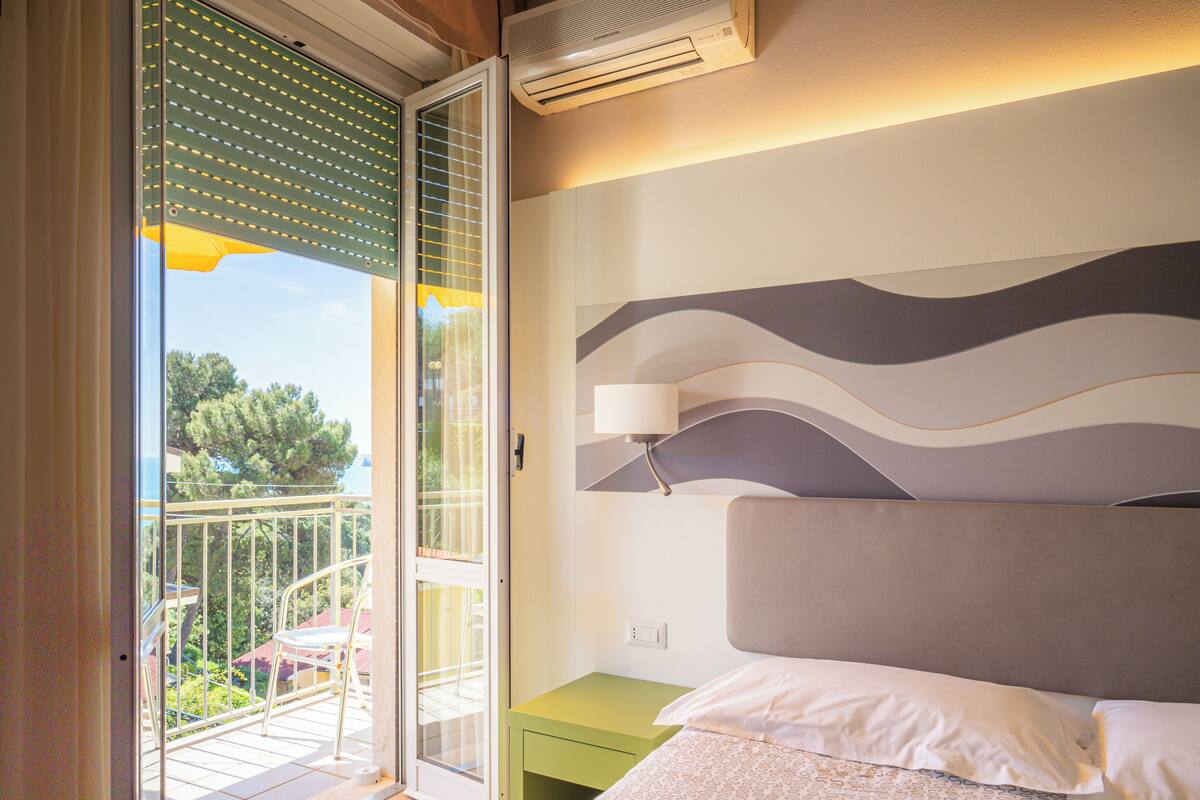 A bedroom with a bed, a nightstand, a wall-mounted lamp, and a balcony with a view of trees and a building.