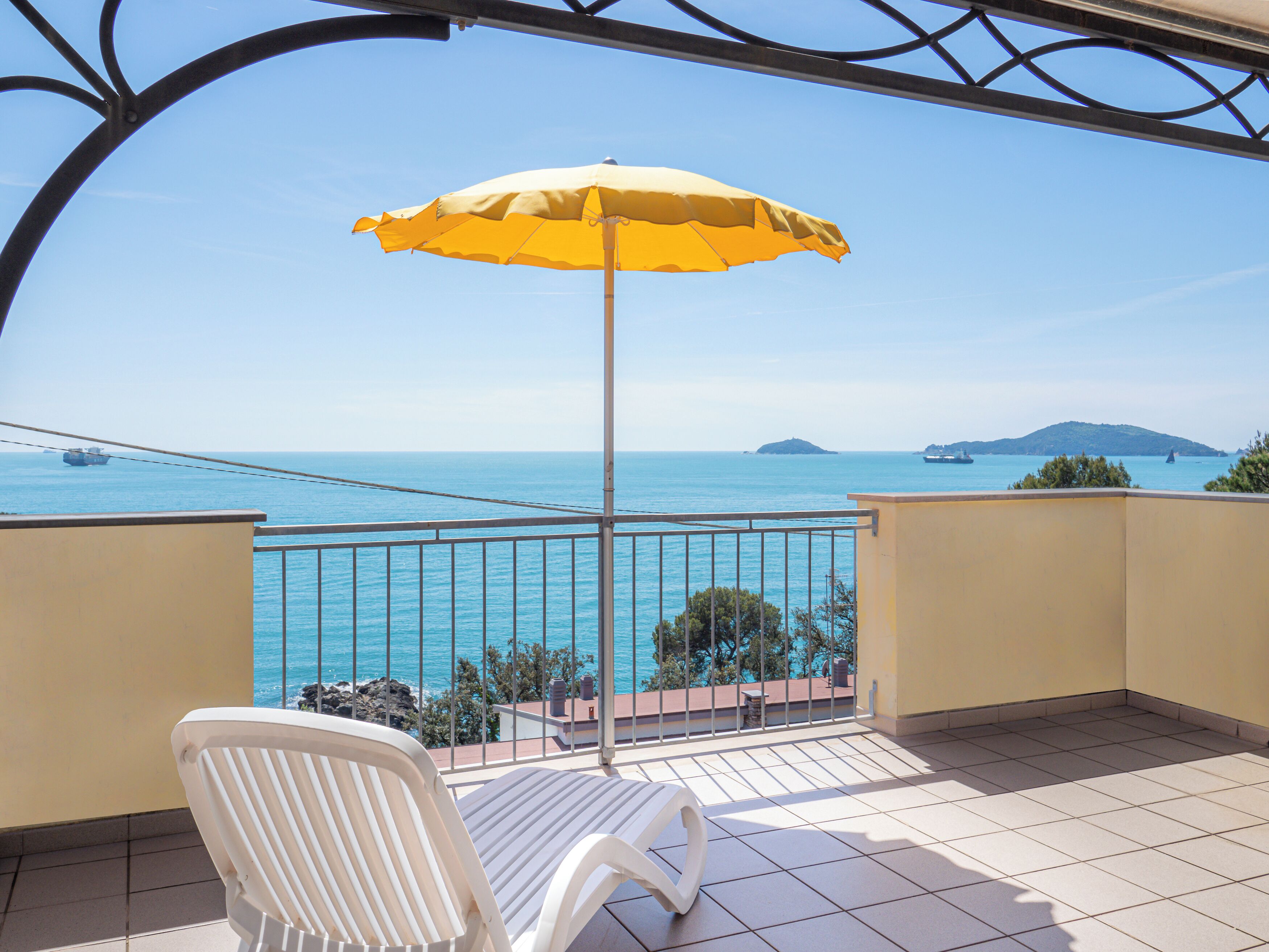 Een balkon met een gele parasol, een witte stoel en uitzicht op zee.