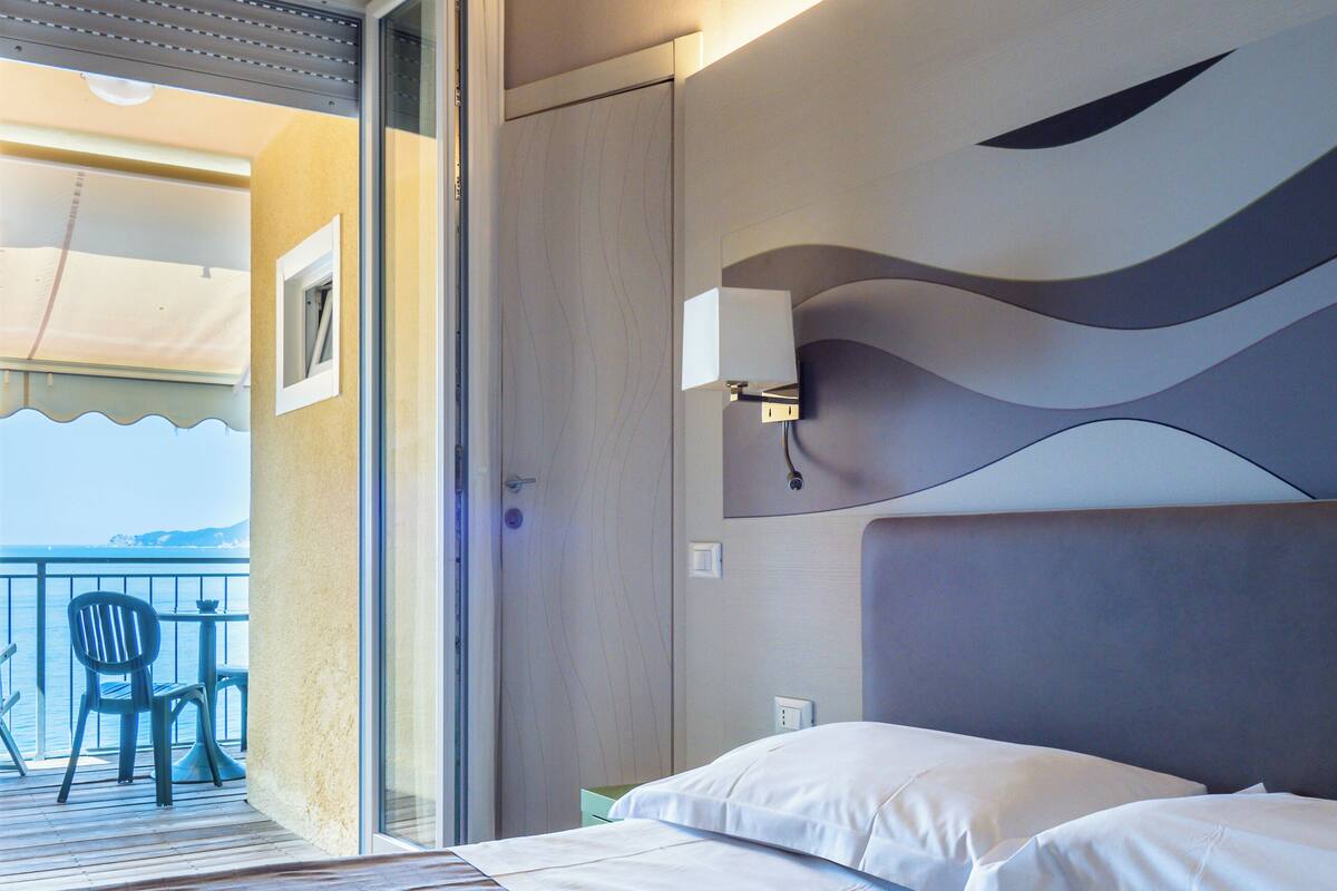A hotel room with a bed, a nightstand, a wall-mounted lamp, a window with curtains, and a view of the sea through the balcony.