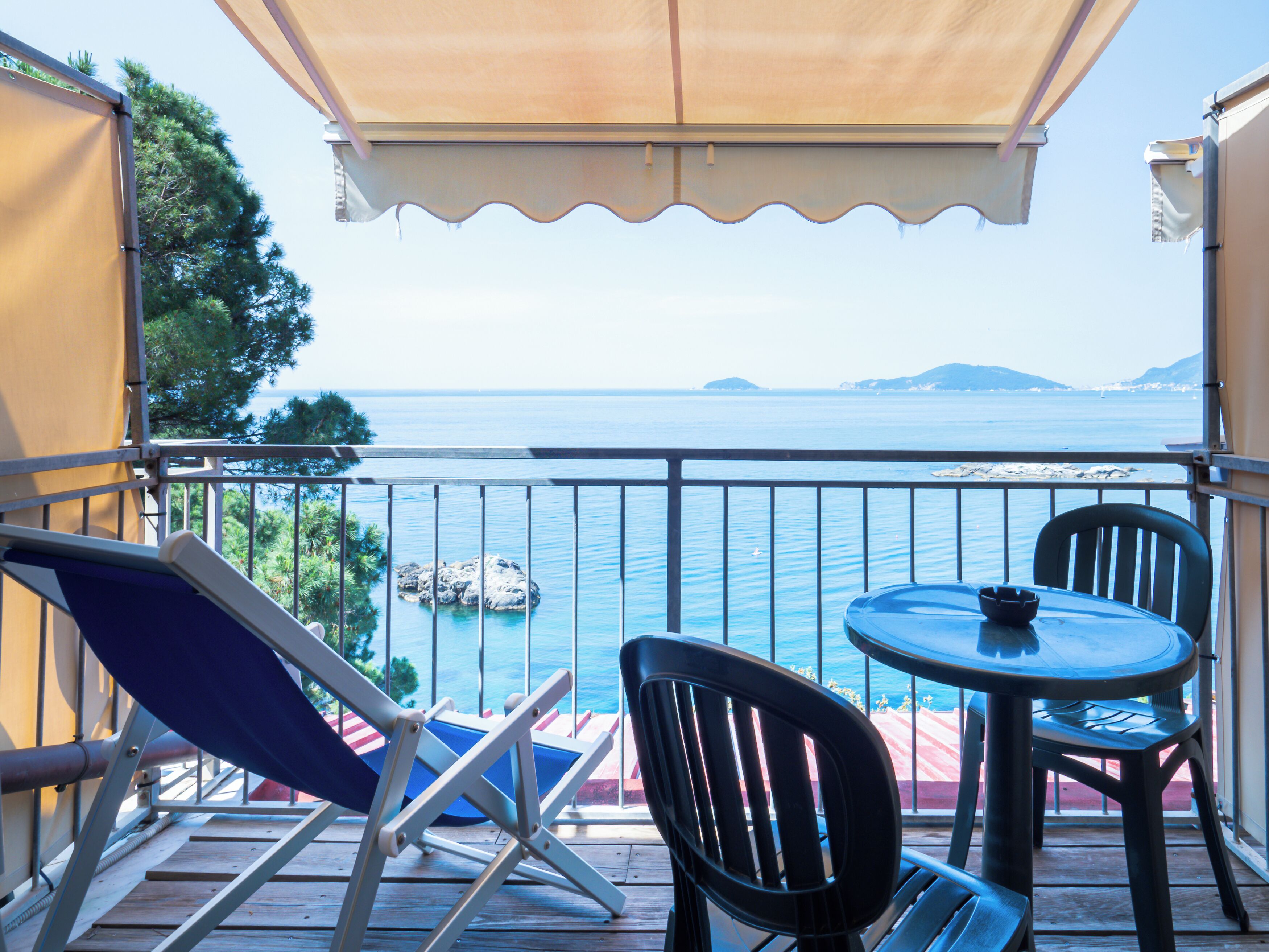 A balcony with a table and chairs, overlooking a body of water with a rocky outcrop and distant landforms.