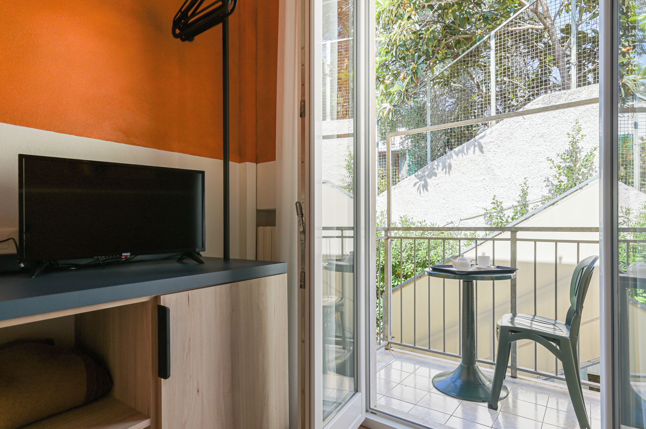 A room with a TV on a wooden cabinet, a balcony with a table and chairs, and a view of a staircase and greenery outside.