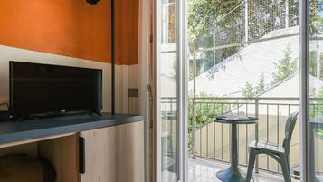 A room with a TV on a wooden cabinet, a balcony with a table and chairs, and a view of a staircase and greenery outside.