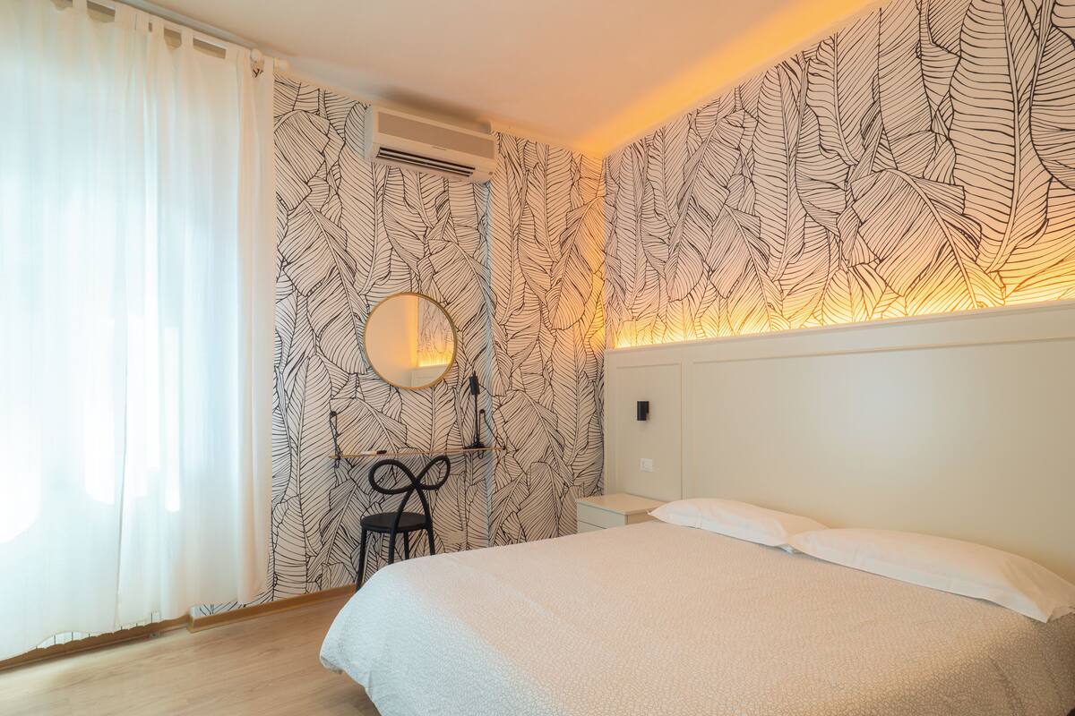 A bedroom with a bed, a desk, a chair, a mirror, and a wall with a leaf pattern.