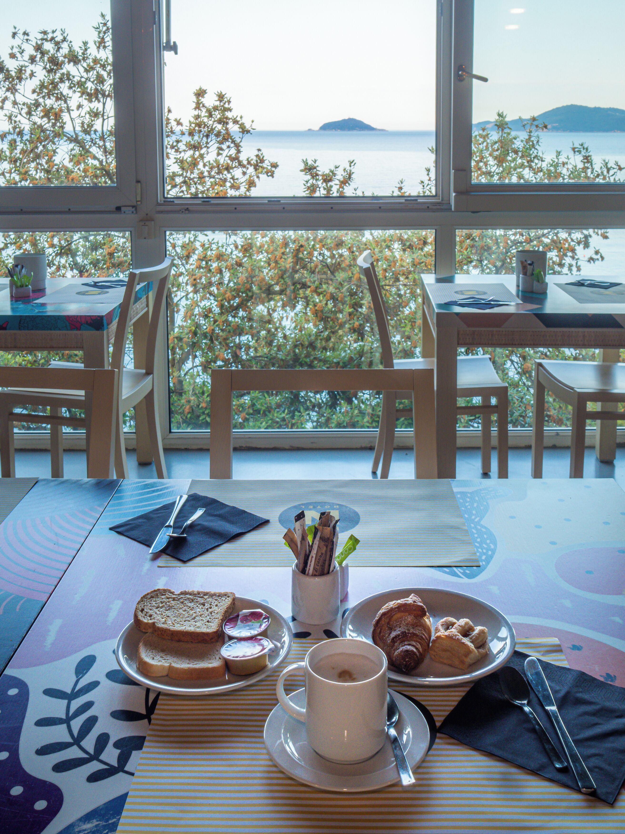 Une table à manger avec des aliments pour le petit-déjeuner, une tasse de café et une vue sur la mer par de grandes fenêtres.