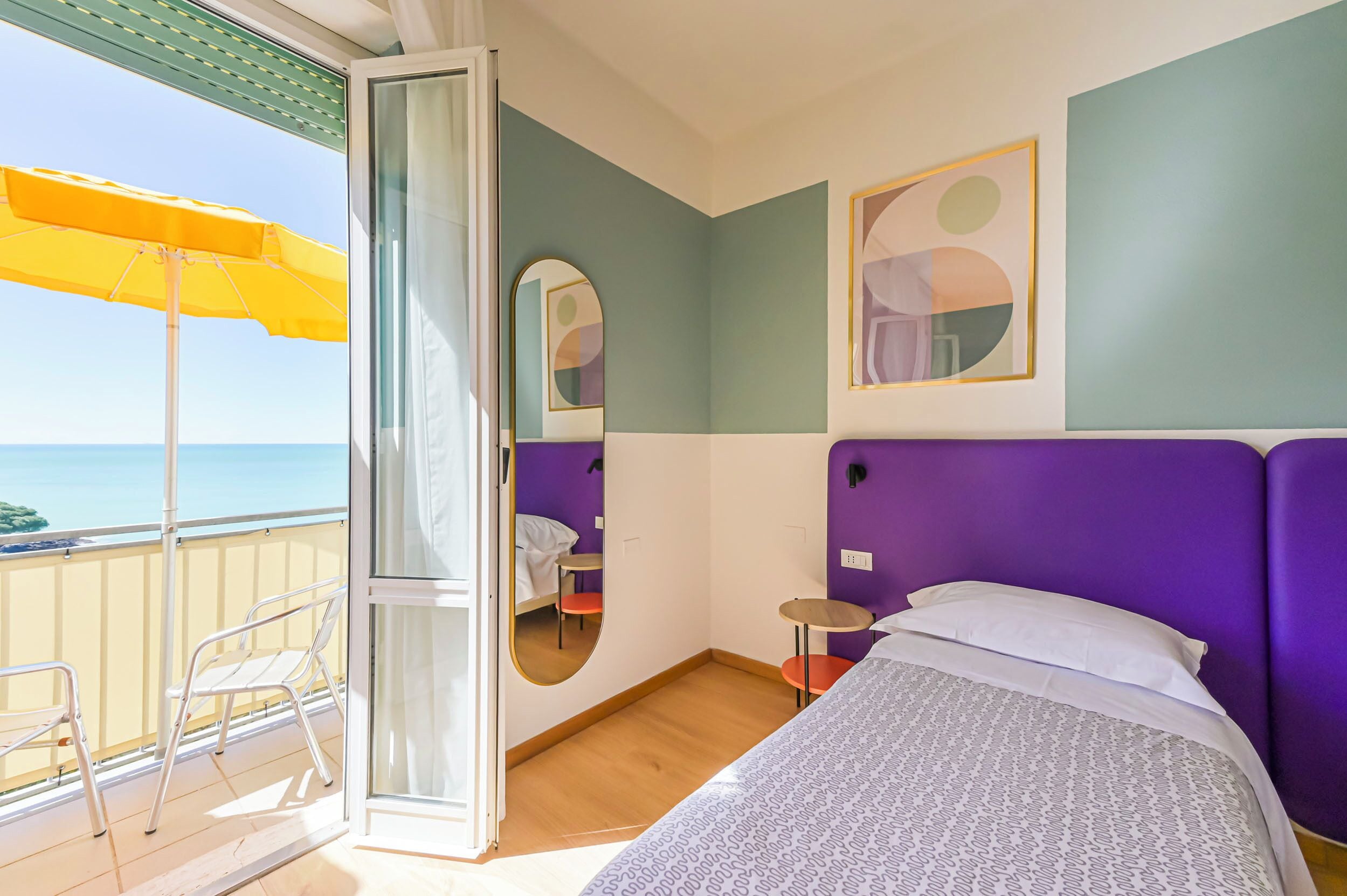 A room with a bed, a mirror, a small table, a chair, and a balcony with an umbrella.
