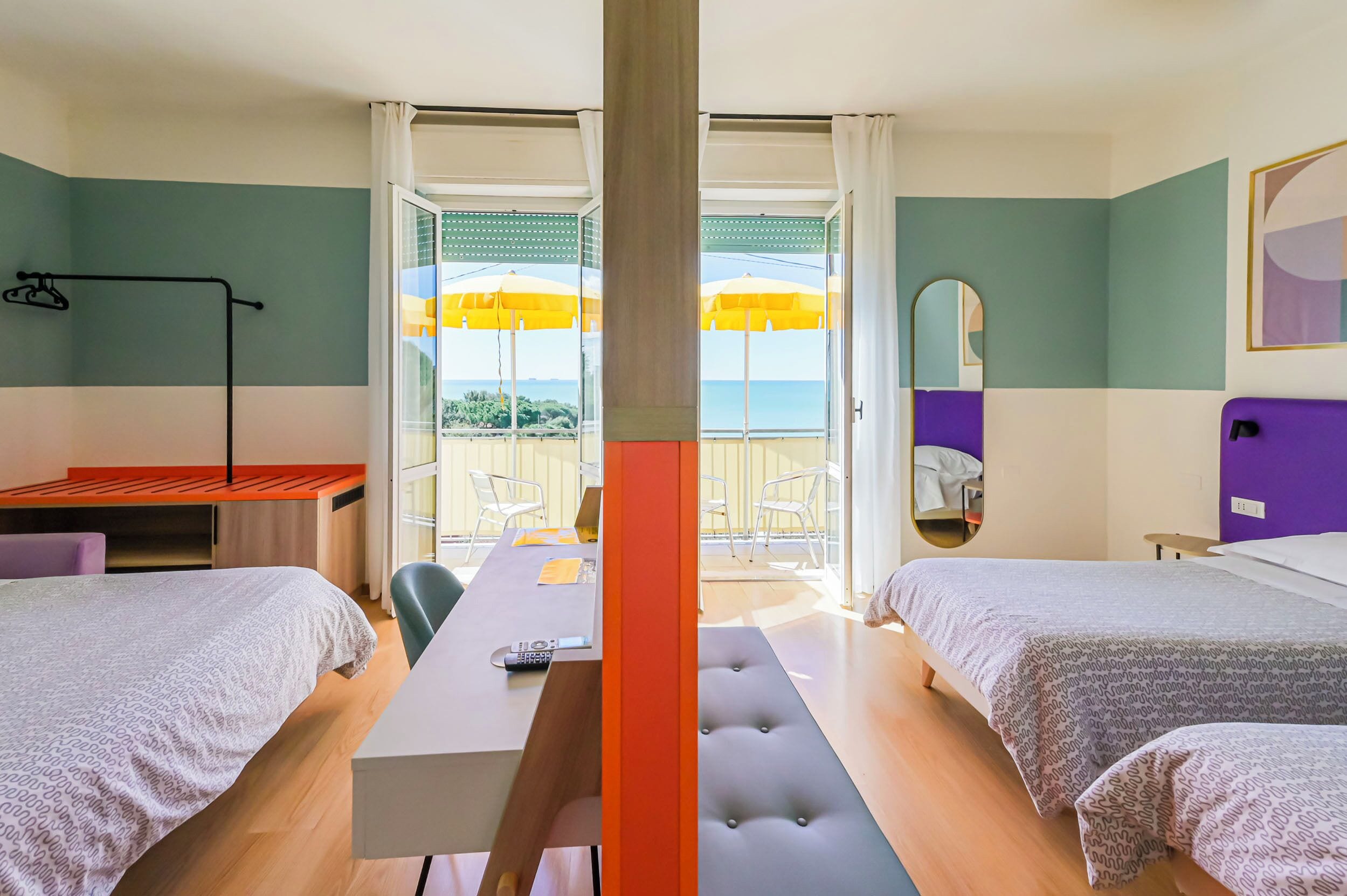 Una camera d'albergo con due letti, una scrivania, una sedia, un balcone con ombrelloni gialli e vista sul mare.
