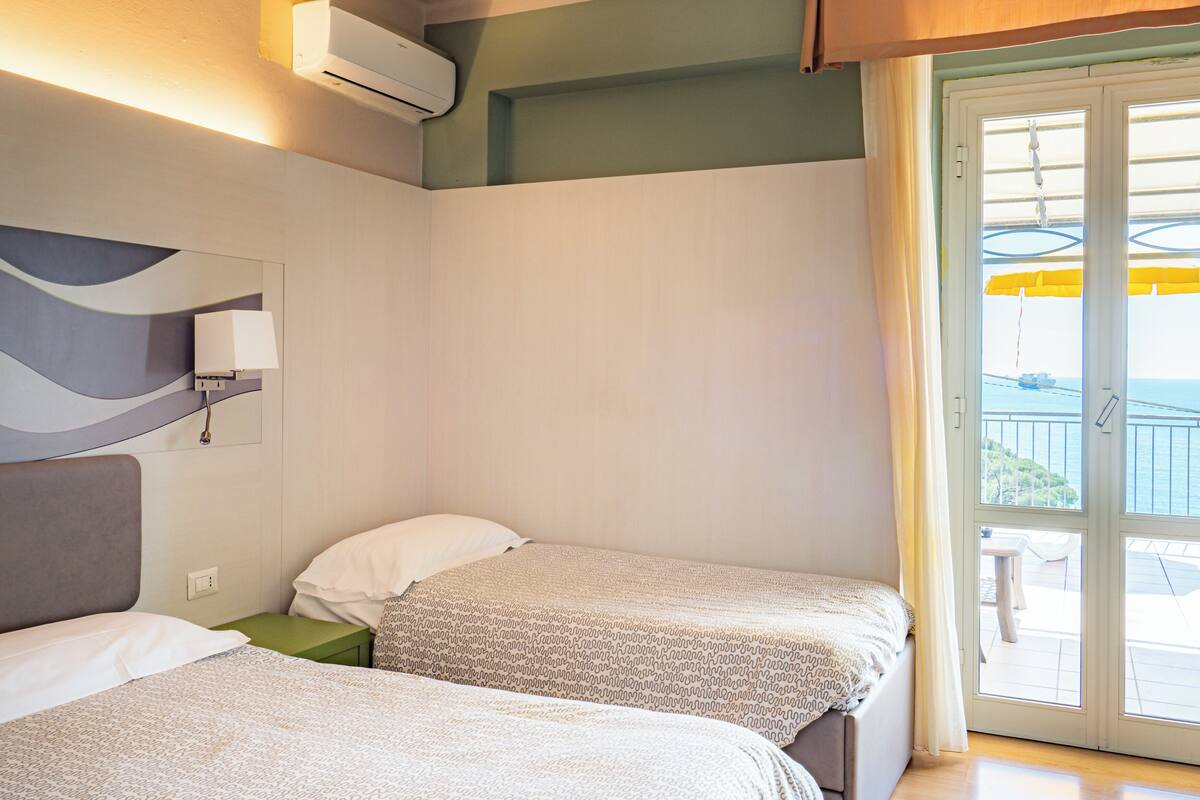 A hotel room with two beds, a window with a view of the sea, and a balcony with yellow umbrellas.