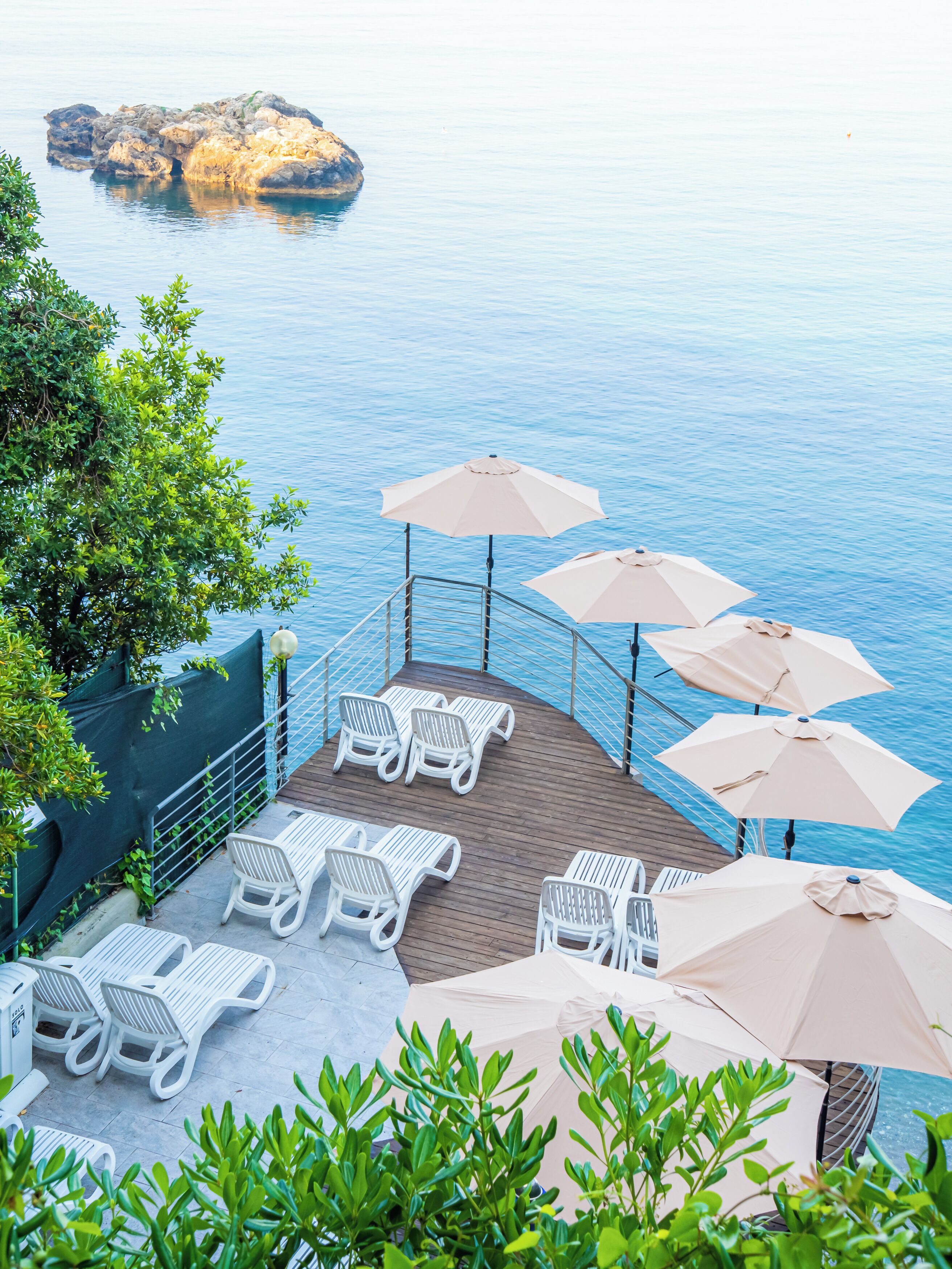 Een terras met witte loungestoelen en beige parasols met uitzicht op een waterpartij met een rotsachtige richel in de verte.