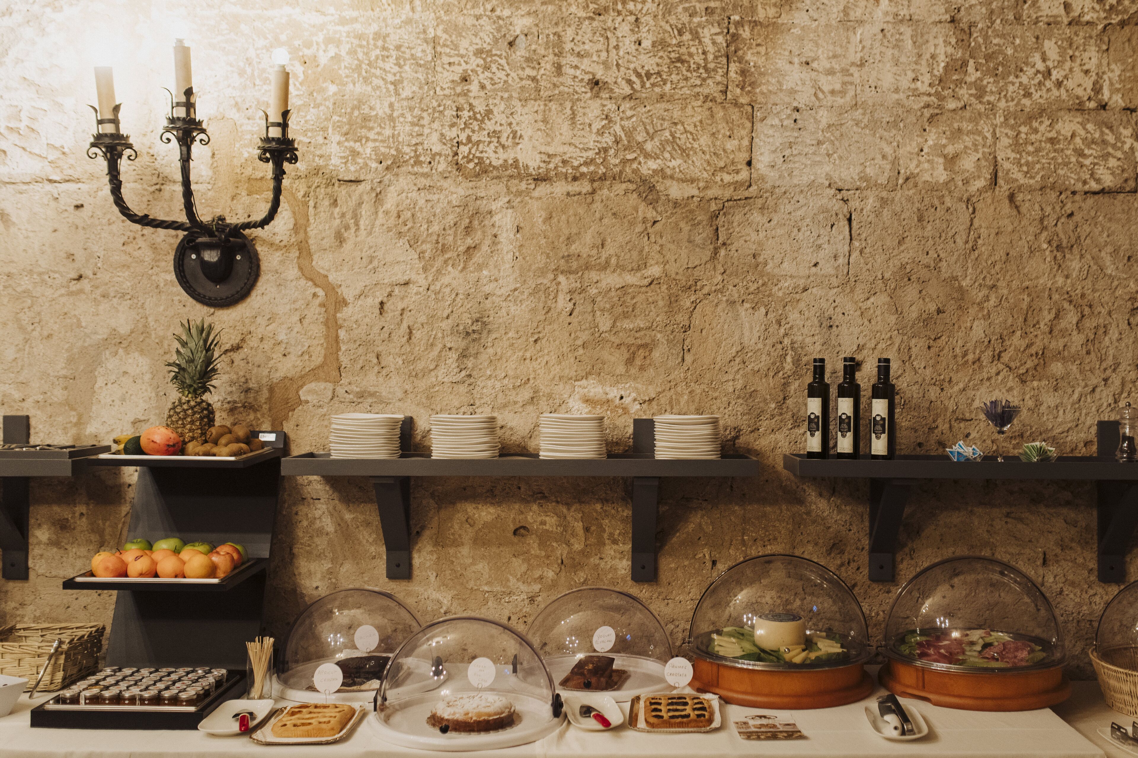 A buffet setup with various food items, including fruits, pastries, and covered dishes, displayed on shelves and tables against a textured wall.