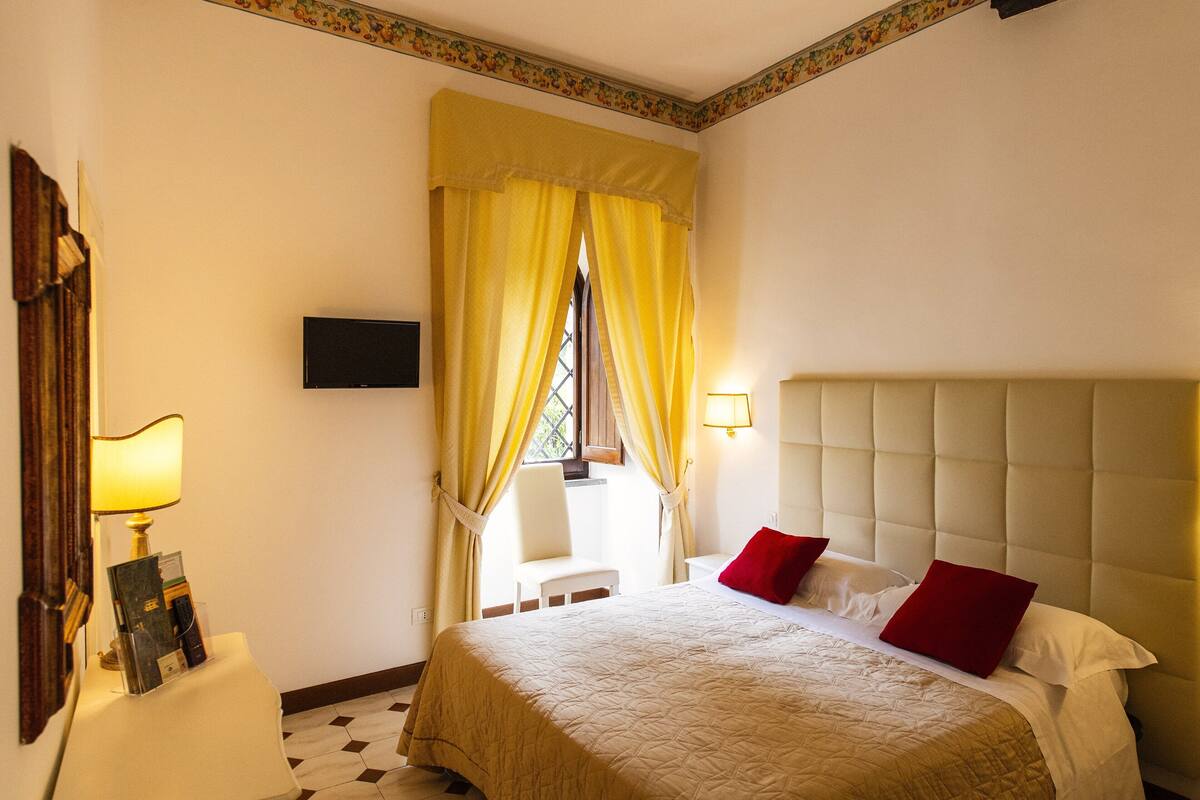 A hotel room with a bed, a bedside table with a lamp, a chair, and a window with curtains.