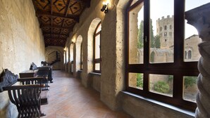 A corridor with stone walls, wooden benches, and large windows overlooking a castle.