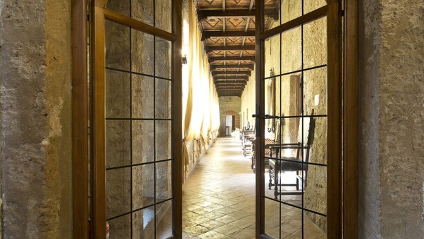 A corridor with arched wooden doors and a tiled floor, leading to a room with a bed and a window.