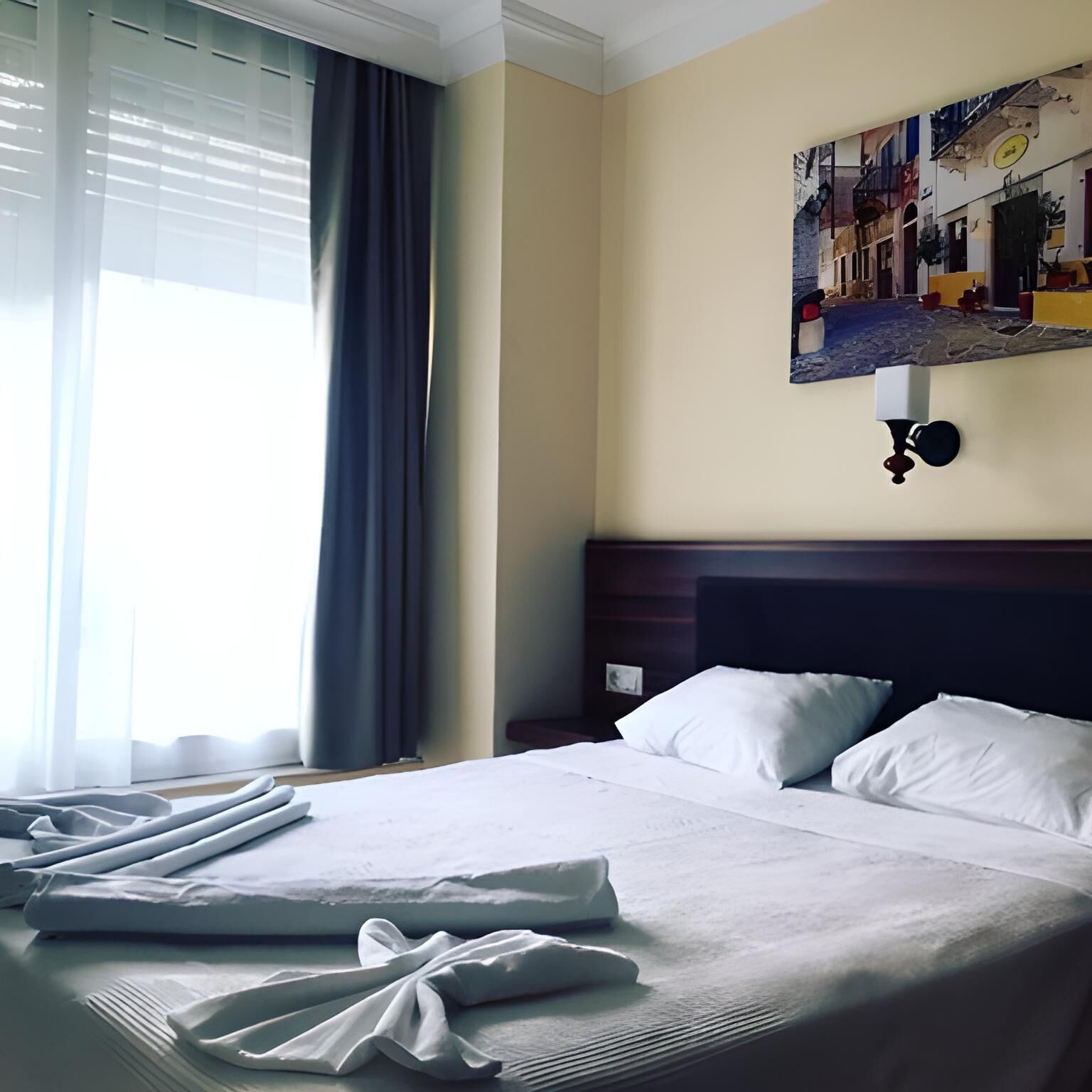 A hotel room with a neatly made bed, a nightstand, a wall-mounted lamp, and a window with curtains.