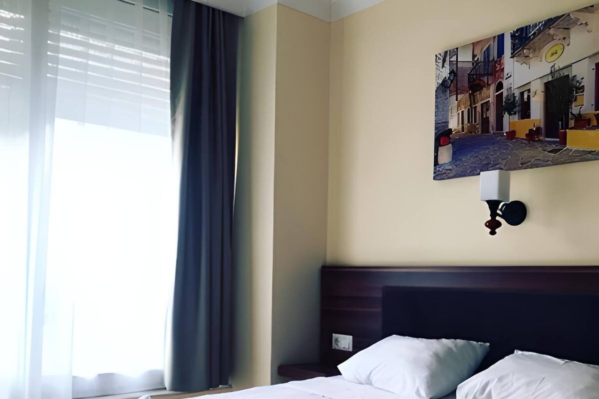 A hotel room with a neatly made bed, a nightstand, a wall-mounted lamp, and a window with curtains.