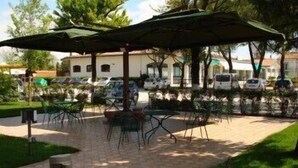 Ein Außenbereich mit Terrasse, Tischen und Stühlen unter Sonnenschirmen, umgeben von Grünflächen und parkenden Autos.