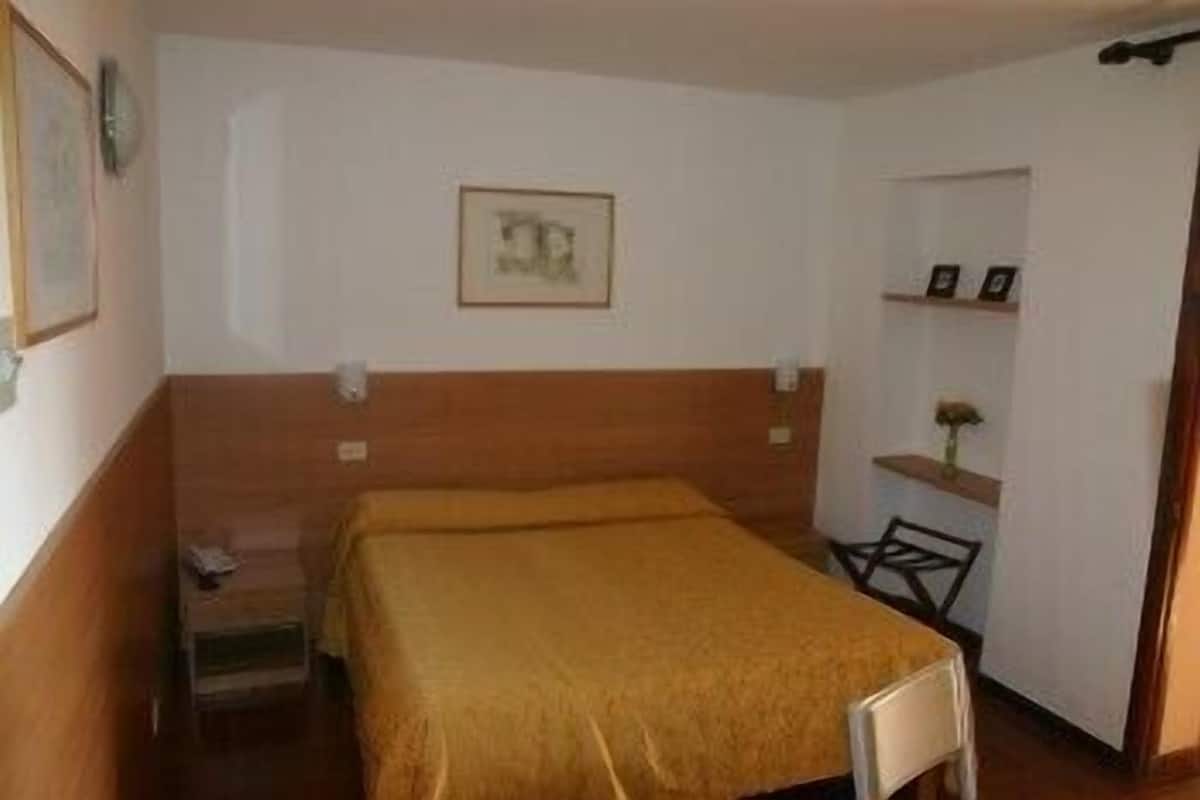 Una habitación pequeña con una cama individual, cabecera de madera, una mesita de noche y un estante con un cuadro enmarcado y un florero.