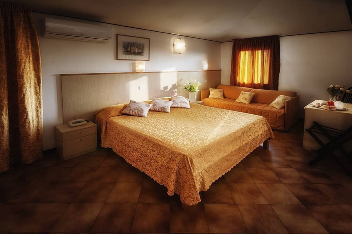 Habitación de hotel con una cama grande, un sofá más pequeño, una mesita de noche con lámpara y una ventana con cortinas.