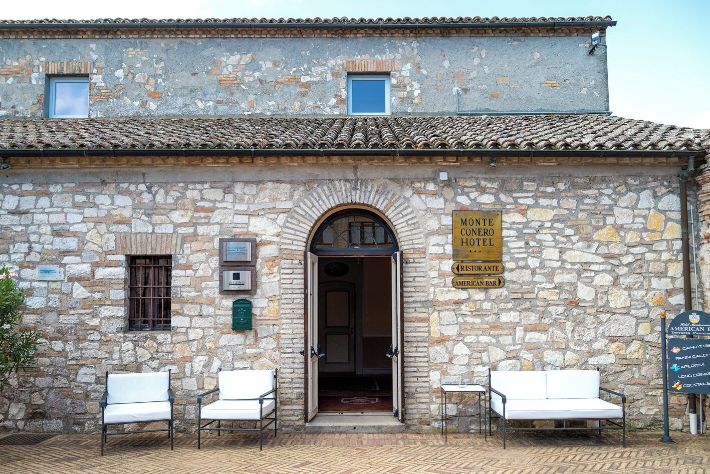 Un bâtiment en pierre avec une enseigne indiquant l’hôtel Monte Conero, un restaurant et un bar à l’américaine. Deux chaises blanches sont placées à l’extérieur.