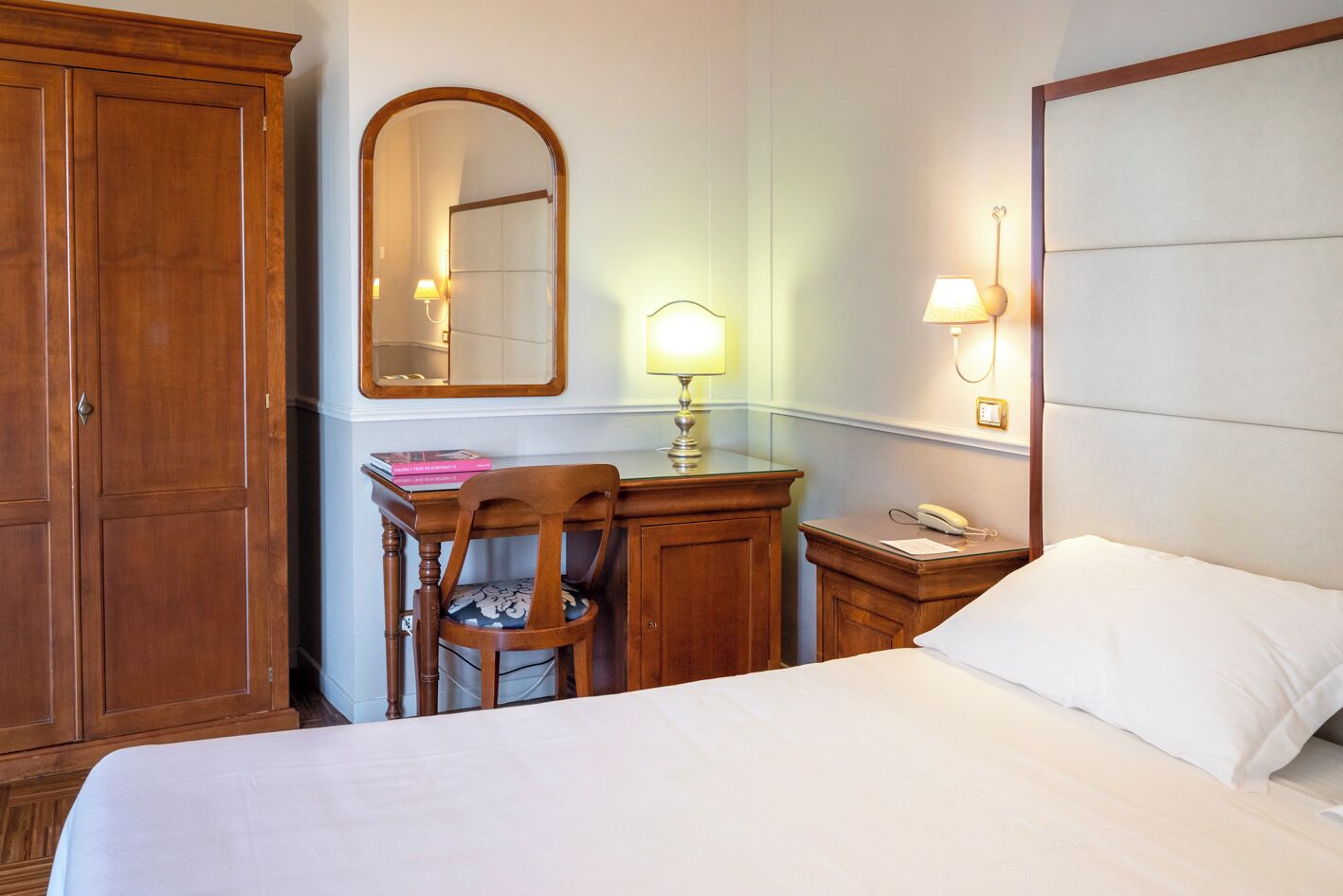 A hotel room with a large bed, a wooden desk, a chair, a mirror, a lamp, and a bedside table with a phone.