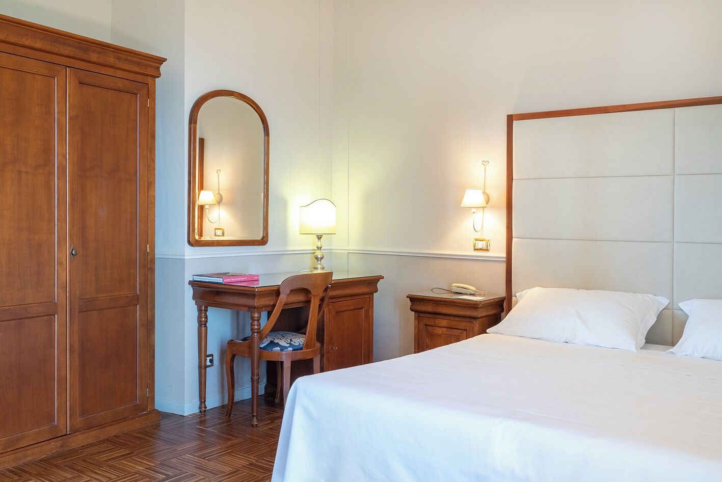 Una camera d'albergo con un letto grande, una scrivania con sedia, un armadio, uno specchio e lampade ai comodini.