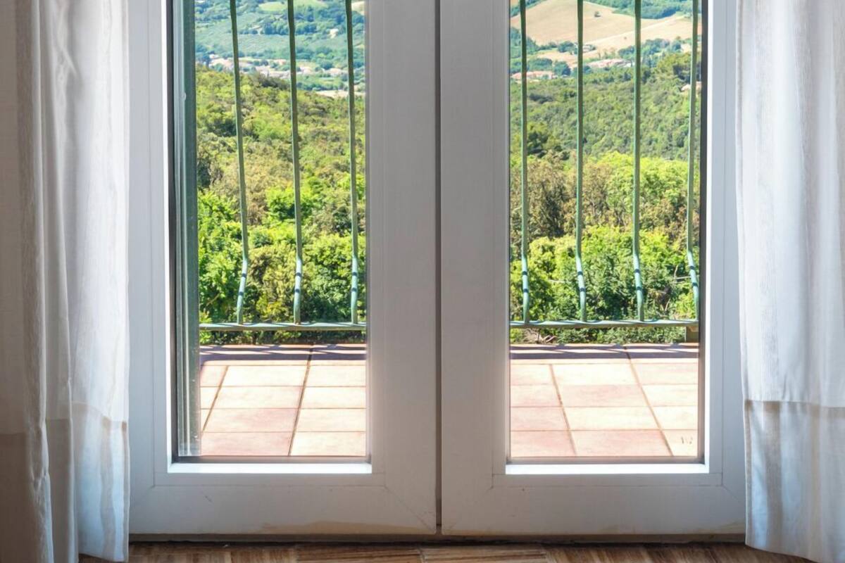 Ein Zimmer mit großem Fenster und Blick auf einen Balkon mit Metallgeländer, viel Grün und ein fernes Gebirge.