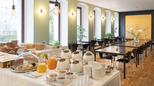 A breakfast buffet with various food items and beverages, a long dining table with chairs, and a large window with natural light. - Hotel Johann (Berlin)