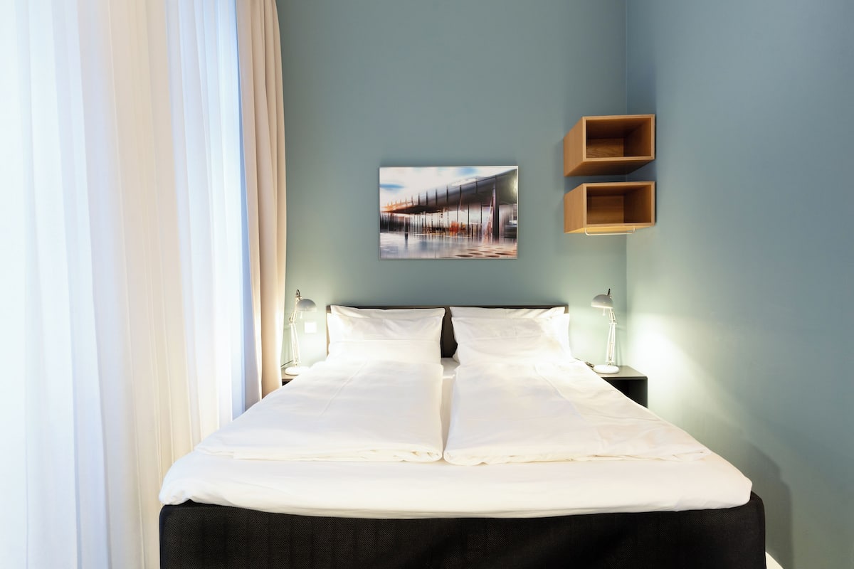 A neatly made bed in a room with a light blue wall, a framed picture, and two wooden shelves.