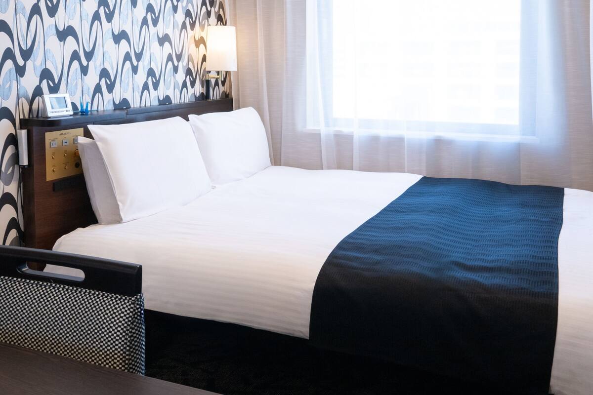 A hotel room with a neatly made bed, a bedside table with a lamp, and a wall clock.